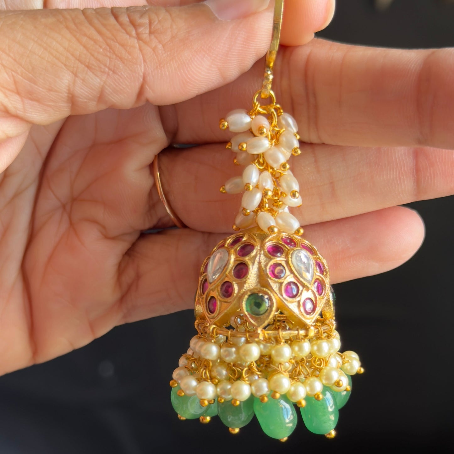A hand holding a gold-toned traditional Indian jhumka earring decorated with white pearls, green stones, and red and purple enamel details.