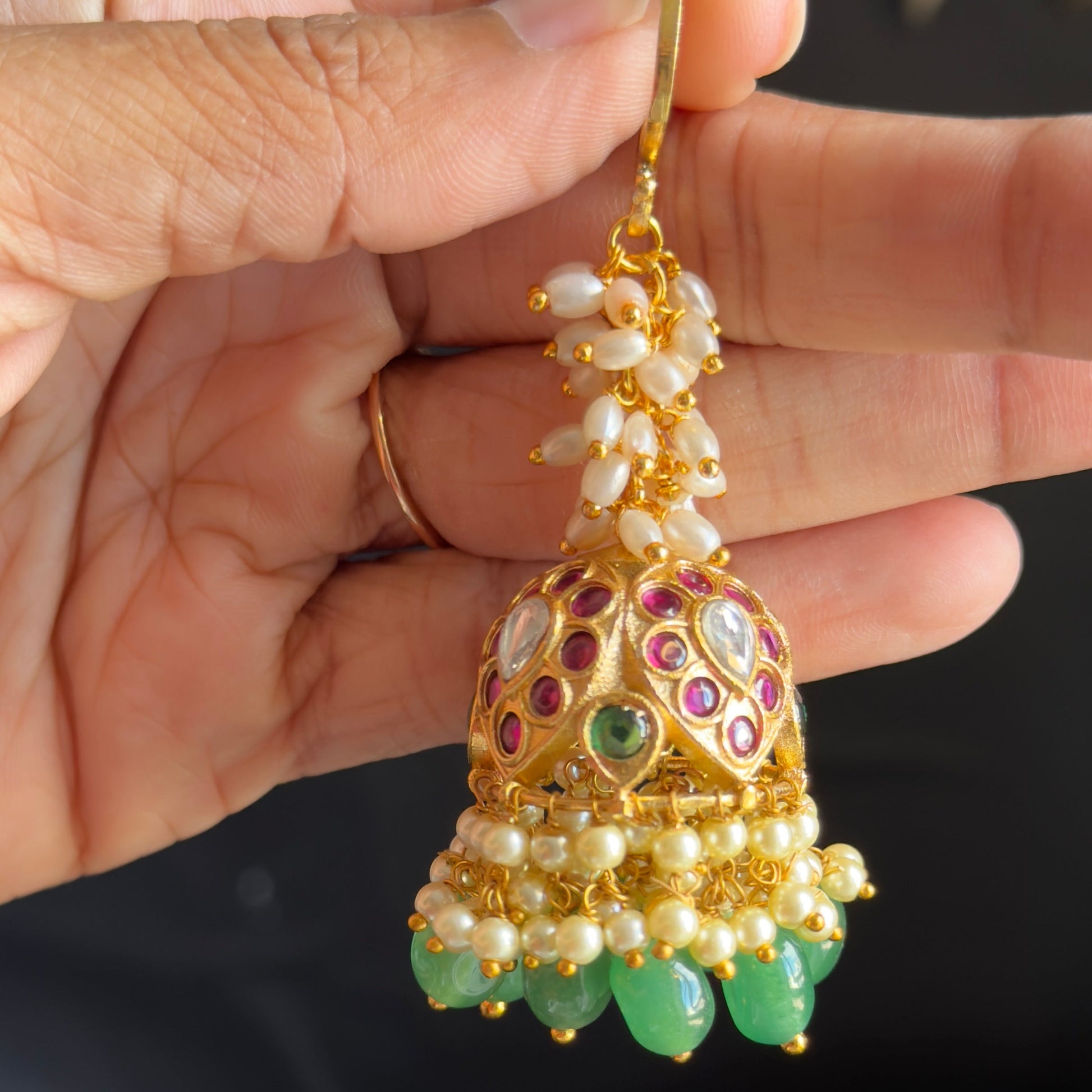 A hand holding a gold-toned traditional Indian jhumka earring decorated with white pearls, green stones, and red and purple enamel details.