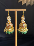 A pair of ornate gold-toned jhumka earrings with white pearl-like beads, multicolored enamel detailing, and dangling green stone accents, displayed on a gold stand against a dark background.