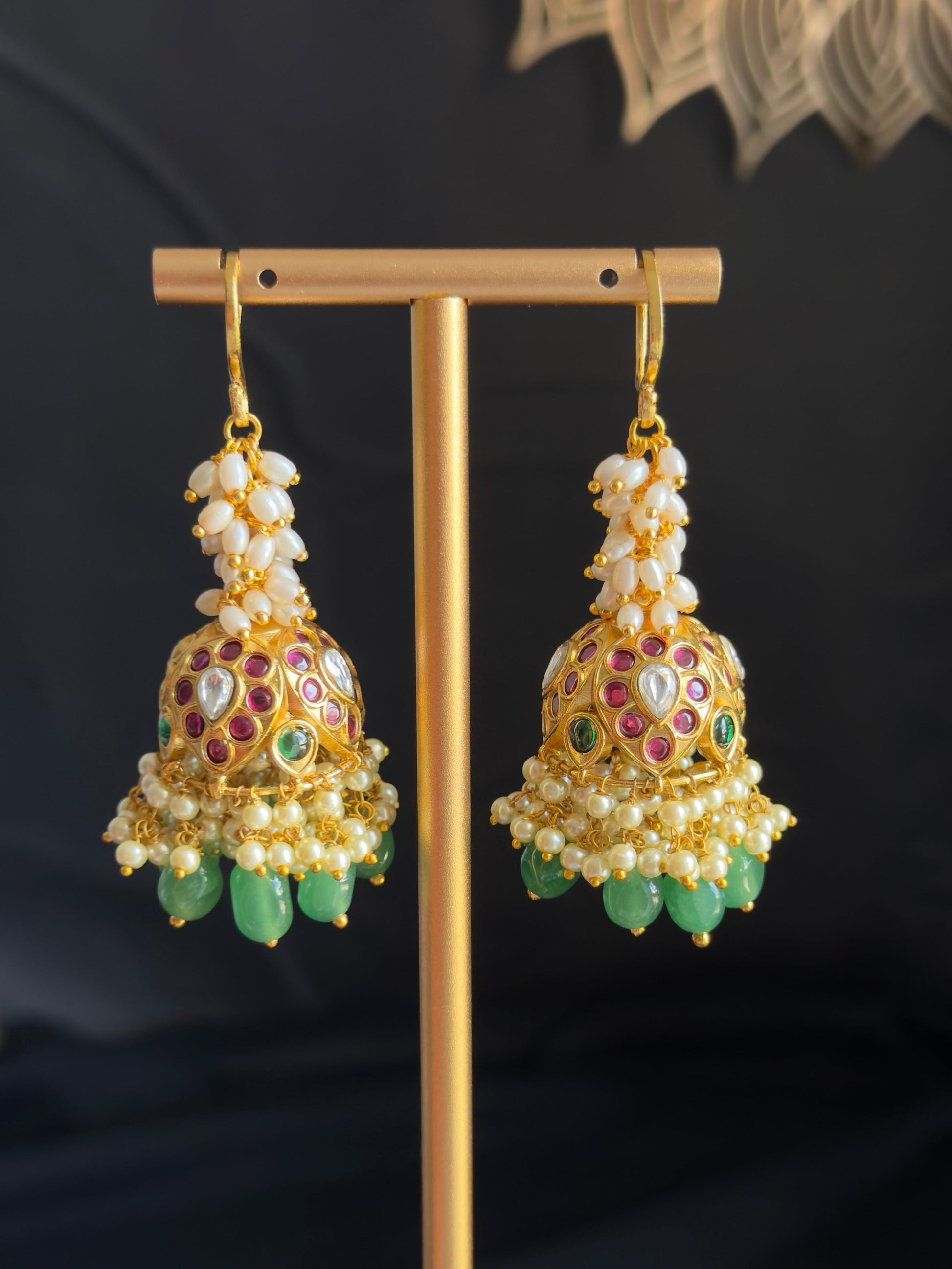 A pair of ornate gold-toned jhumka earrings with white pearl-like beads, multicolored enamel detailing, and dangling green stone accents, displayed on a gold stand against a dark background.