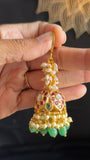A hand holding a gold-toned traditional Indian jhumka earring decorated with white pearls, green stones, and red and purple enamel details.