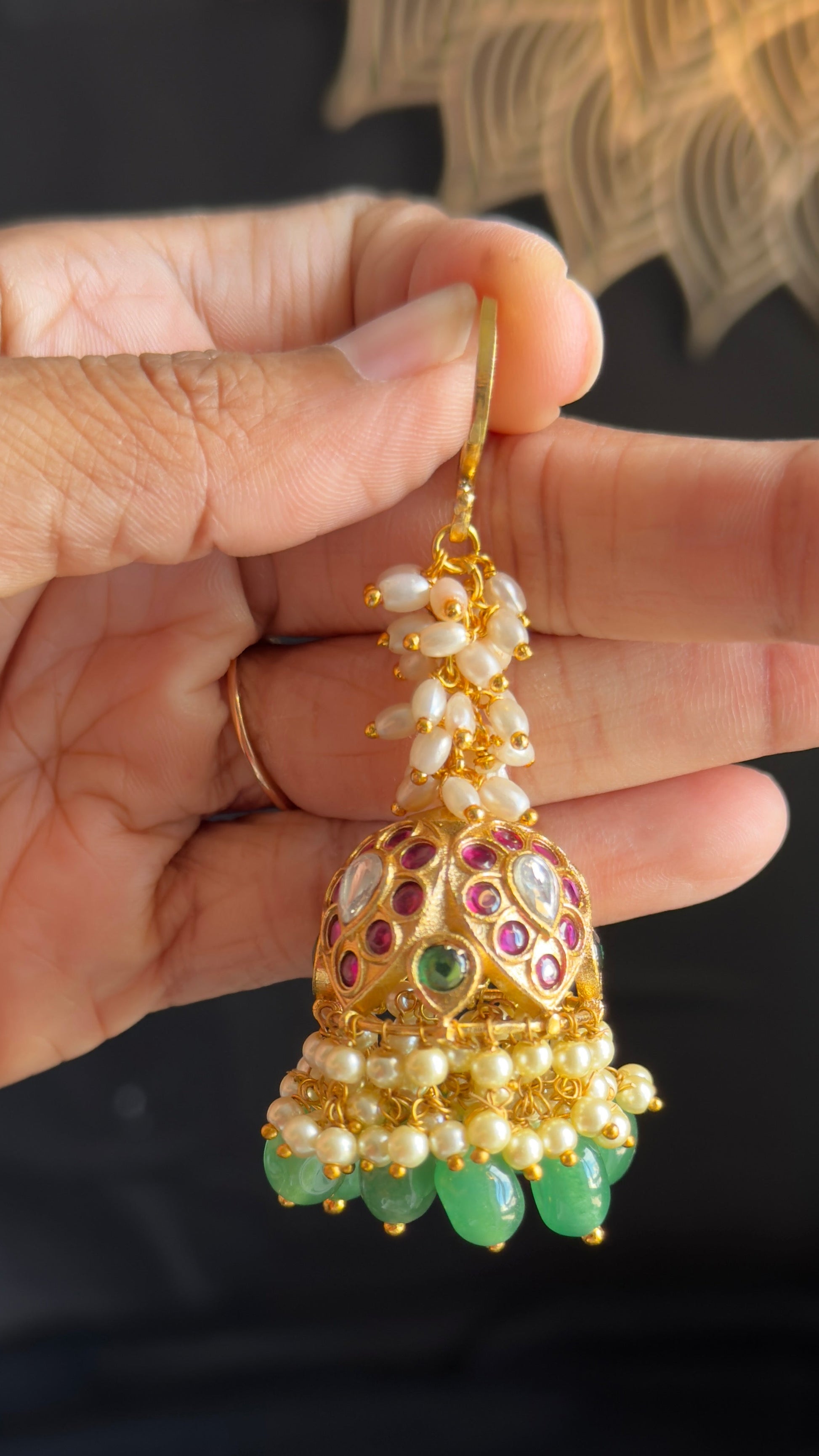 A hand holding a gold-toned traditional Indian jhumka earring decorated with white pearls, green stones, and red and purple enamel details.