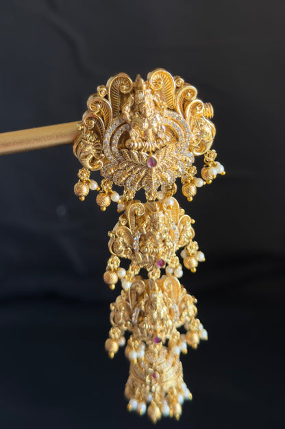 An ornate gold-toned traditional Indian jhumka headpiece with intricate filigree work, beadwork, and dangling elements, held horizontally by a wooden stick against a dark background.
