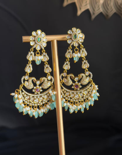 A pair of ornate gold-toned dangle earrings with clear stones, light blue beads, and small pink gemstone accents, displayed on a gold stand against a dark background.