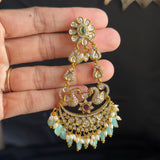 A hand holding a gold-toned traditional Indian jhumka earring decorated with green stones and white bead fringe, set against a dark background.