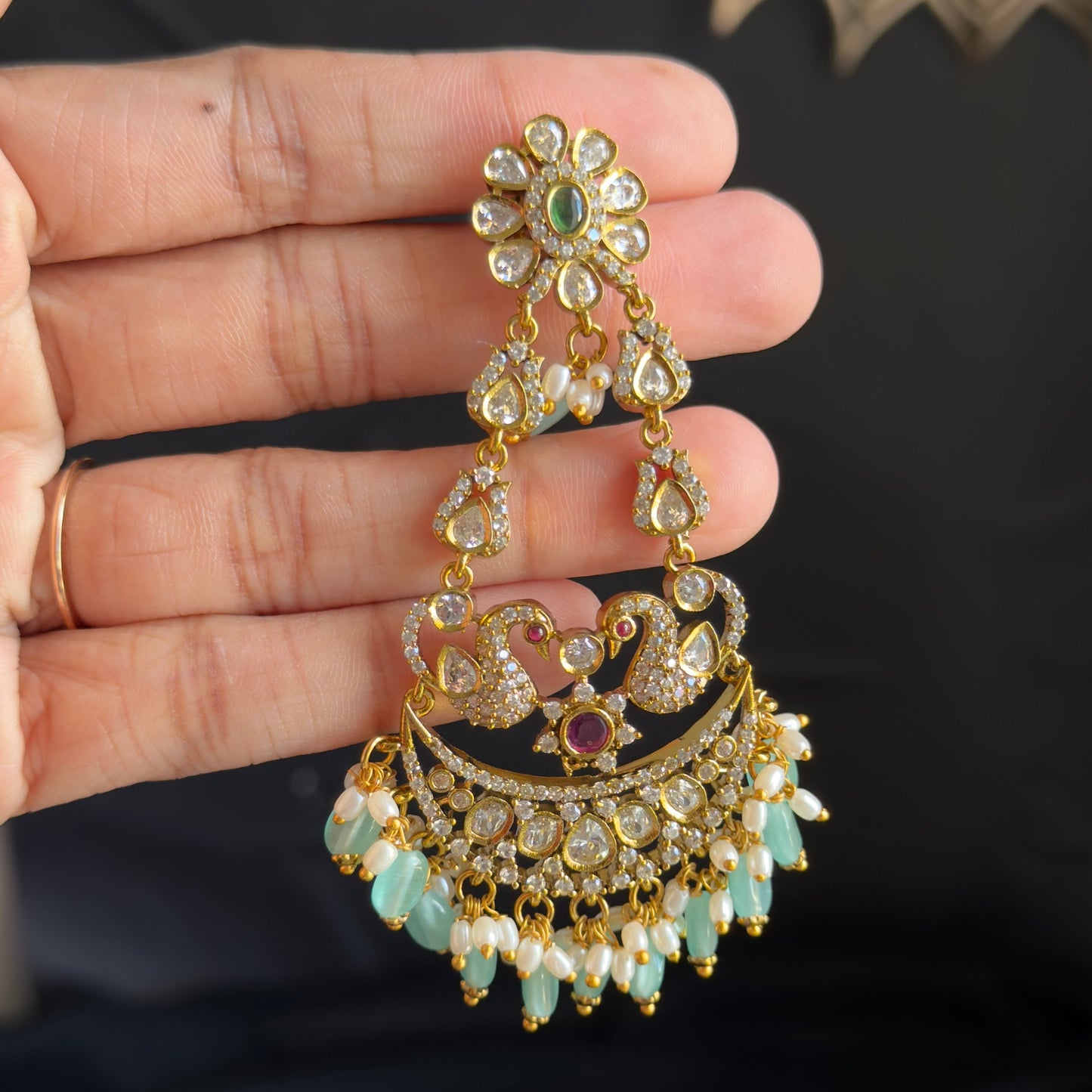 A hand holding a gold-toned traditional Indian jhumka earring decorated with green stones and white bead fringe, set against a dark background.