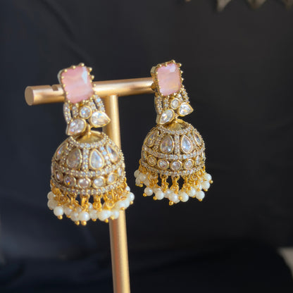 Gold-toned traditional Indian jhumka earrings with pink stones and white pearl fringe, displayed on a gold stand against a dark background.