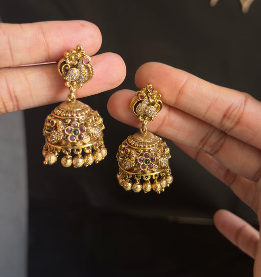 A pair of ornate gold-toned Indian jhumka earrings with colorful enamel detailing, held by two hands against a dark background.