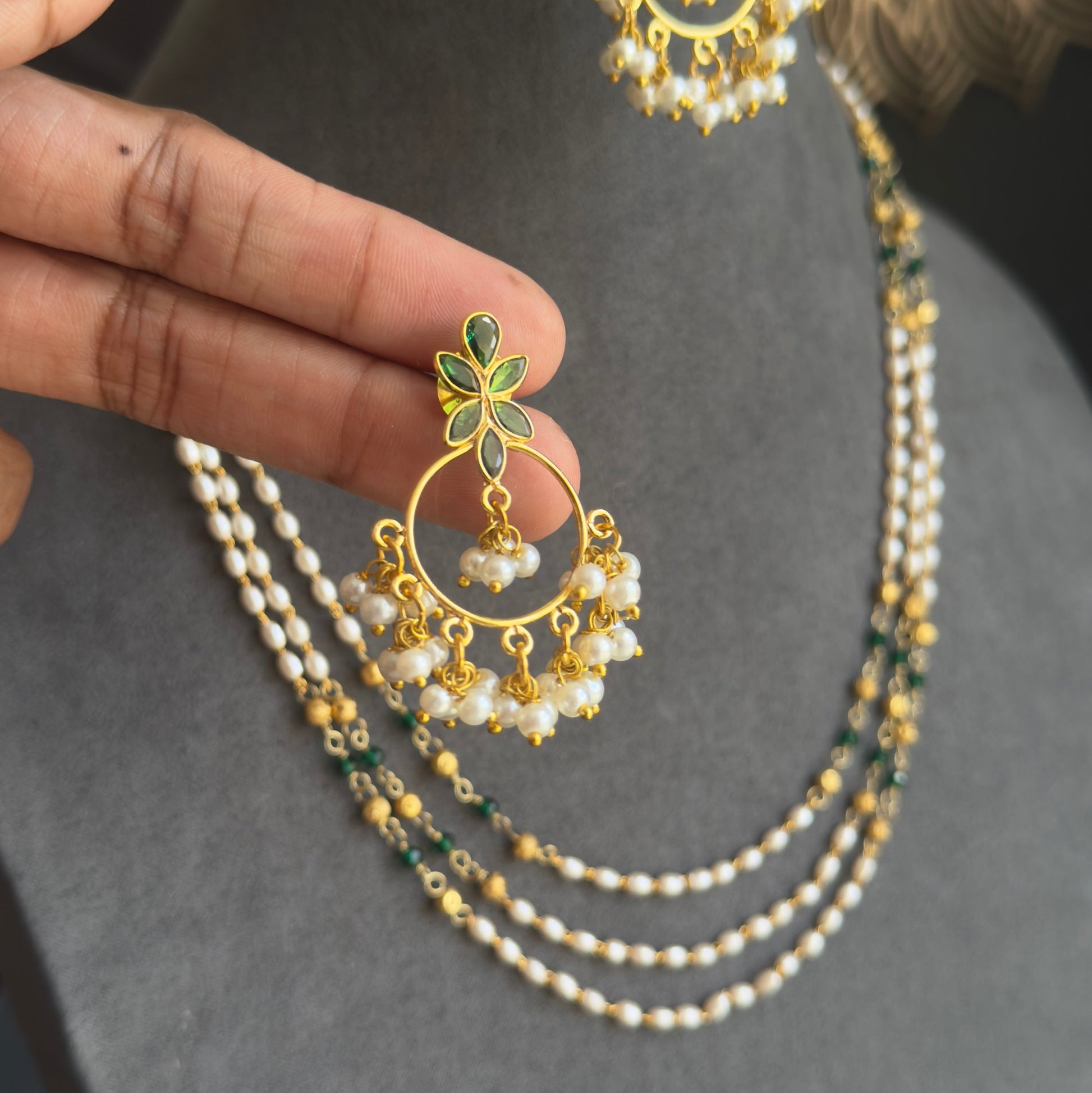 Gold-toned traditional Indian jewelry set featuring a double-strand pearl necklace and matching earrings with green stone accents, displayed on a gray jewelry bust with a hand holding one earring.
