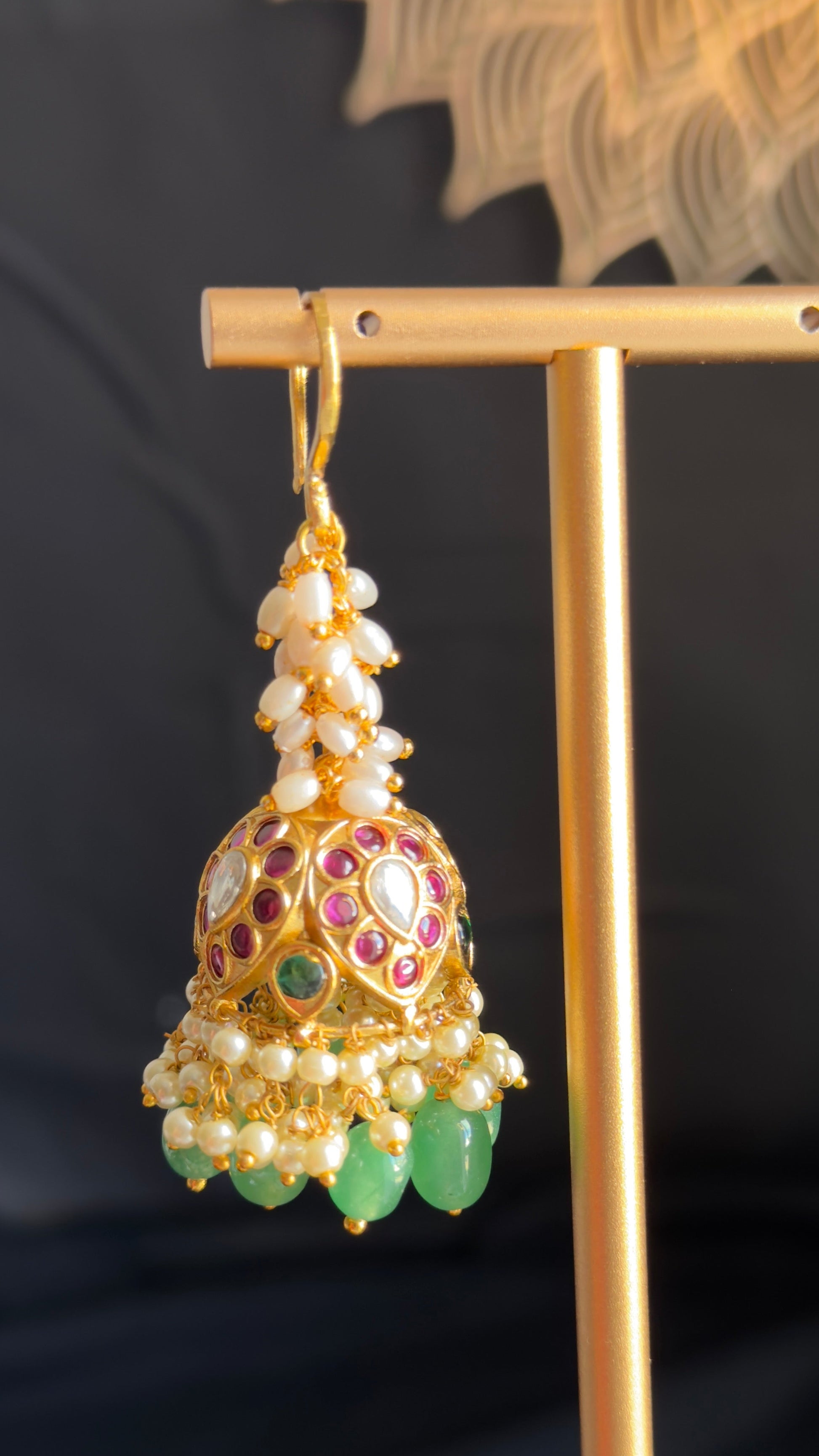 An ornate gold-toned jhumka earring with white pearl-like beads, red and green stone accents, and hanging green teardrop charms, displayed on a gold stand against a dark background.