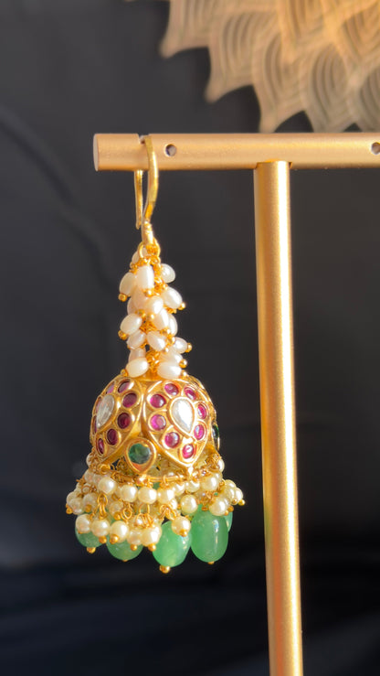 An ornate gold-toned jhumka earring with white pearl-like beads, red and green stone accents, and hanging green teardrop charms, displayed on a gold stand against a dark background.