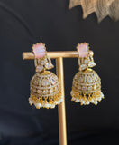 A pair of ornate gold-toned jhumka earrings with pink stones and white pearl bead borders, displayed on a gold stand against a dark background.