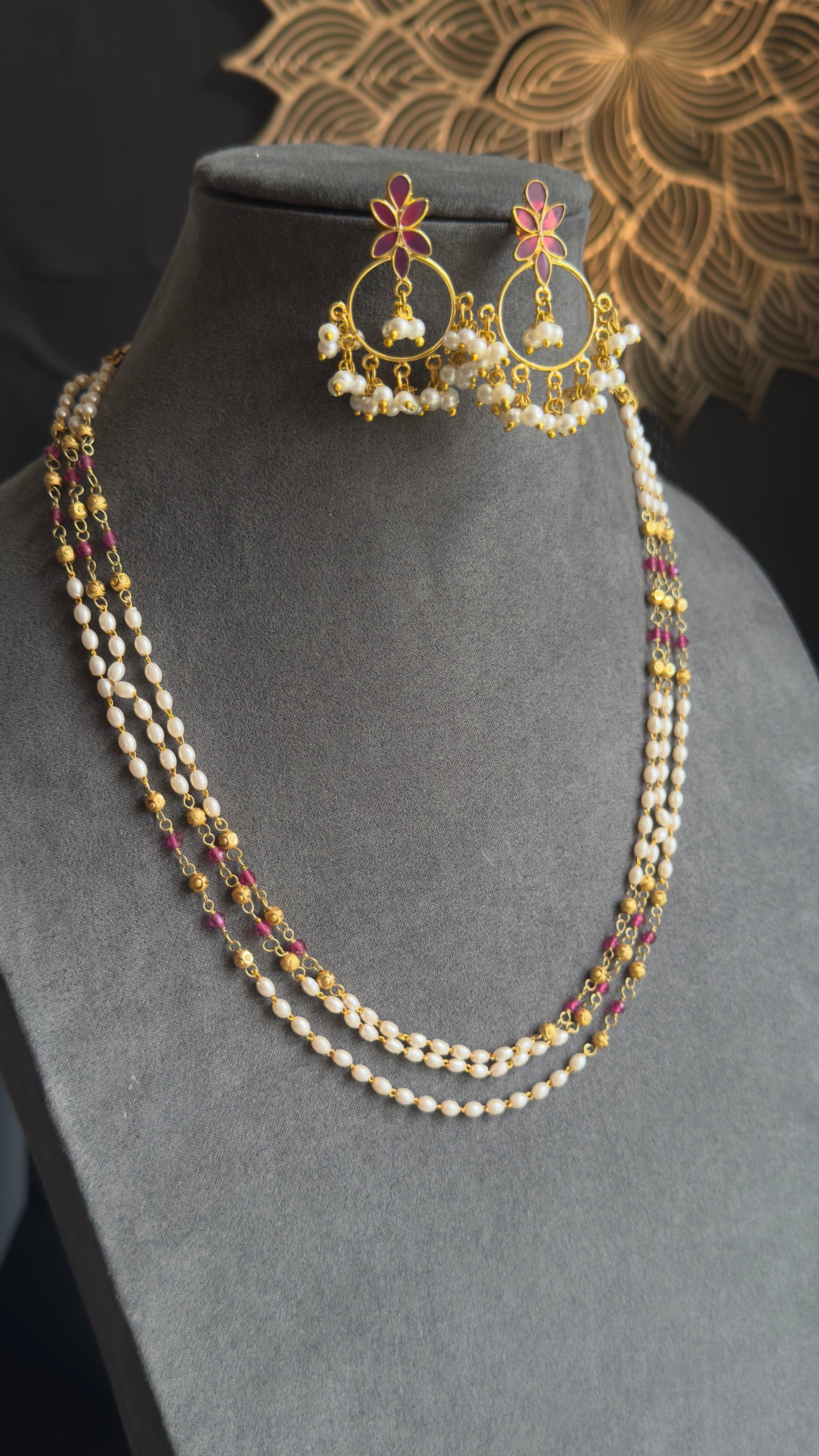 A gold-plated traditional Indian jewelry set featuring a multi-strand necklace with white pearls and red stones, matching drop earrings, and a matching headpiece, displayed on a gray velvet mannequin bust.