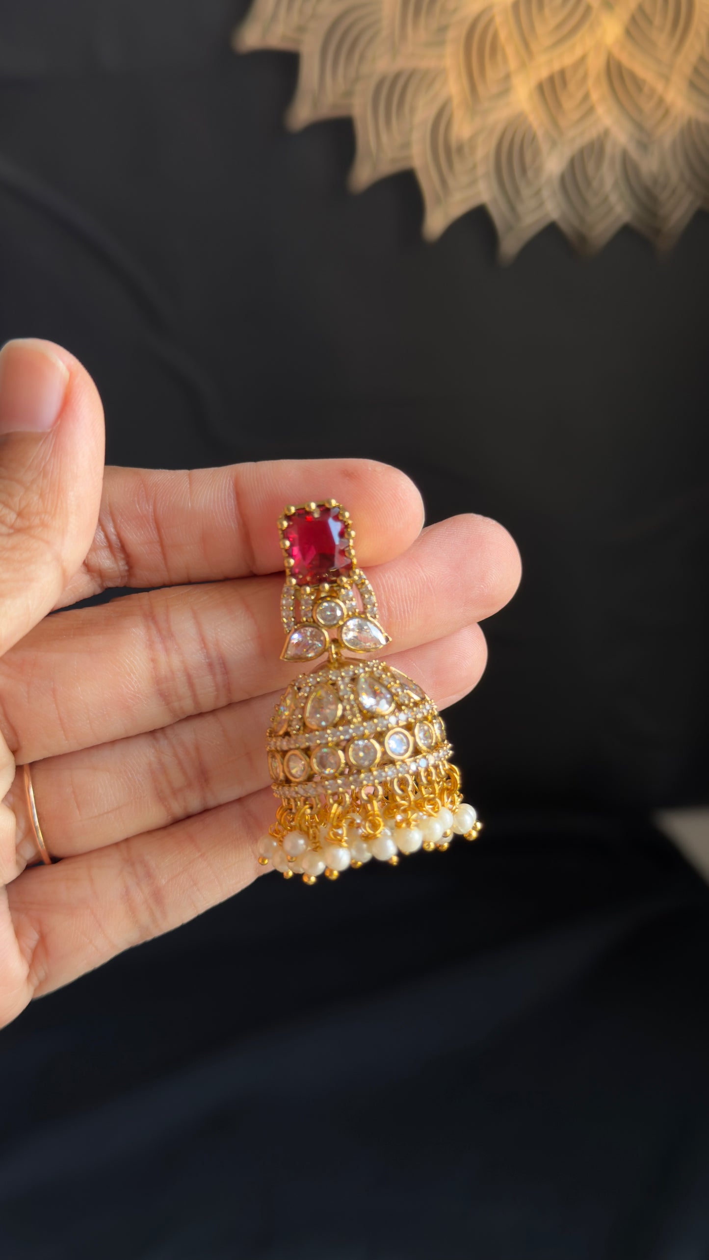A gold-toned traditional Indian jhumka earring with a red gemstone and white pearl accents, held in a person's hand against a dark background.