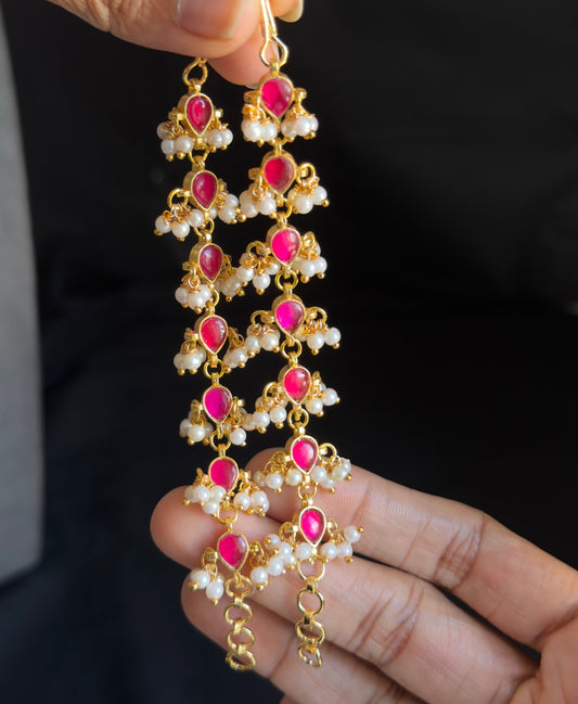A pair of gold-toned traditional Indian jhumka earrings with pink stones and white pearl-like beads, held by a person's hands against a dark background.