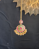 A gold-toned traditional Indian Jhumka pendant necklace with red, green, and gold enamel detailing, held by a hand against a dark background.