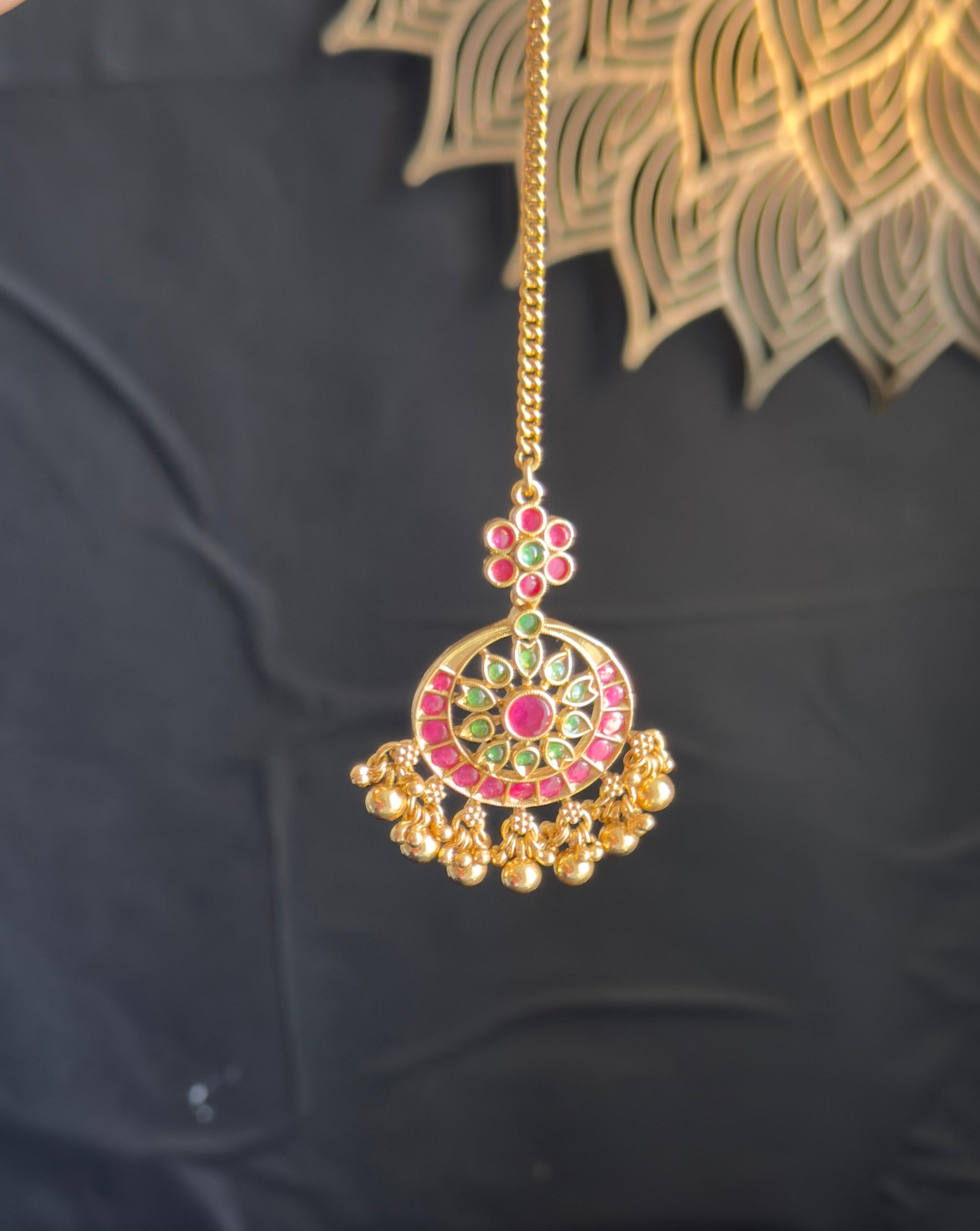 A gold-toned traditional Indian Jhumka pendant necklace with red, green, and gold enamel detailing, held by a hand against a dark background.